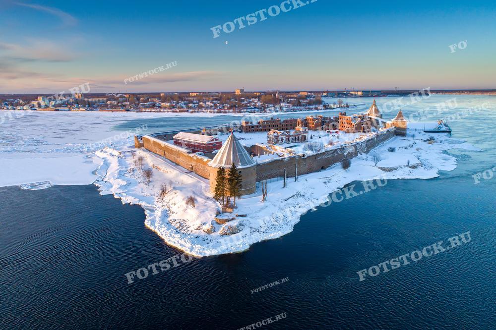 Фотография с высоты демонстрирует крепость Орешек, расположенную на острове в реке Неве в зимнее время. Снег покрывает крепостные стены и землю вокруг, а река покрыта льдом, что создает живописный и мирный пейзаж. Этот снимок идеален для использования в исторических исследованиях, образовательных материалах и публикациях, связанных с историей России.